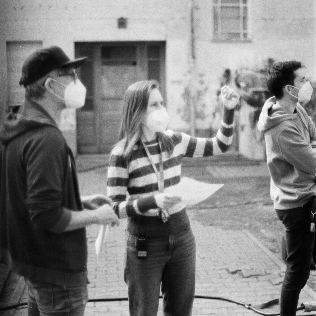 Three people wearing face masks are standing outdoors on a cobblestone surface, engaged in a discussion while holding papers and pointing, with a building facade in the background.