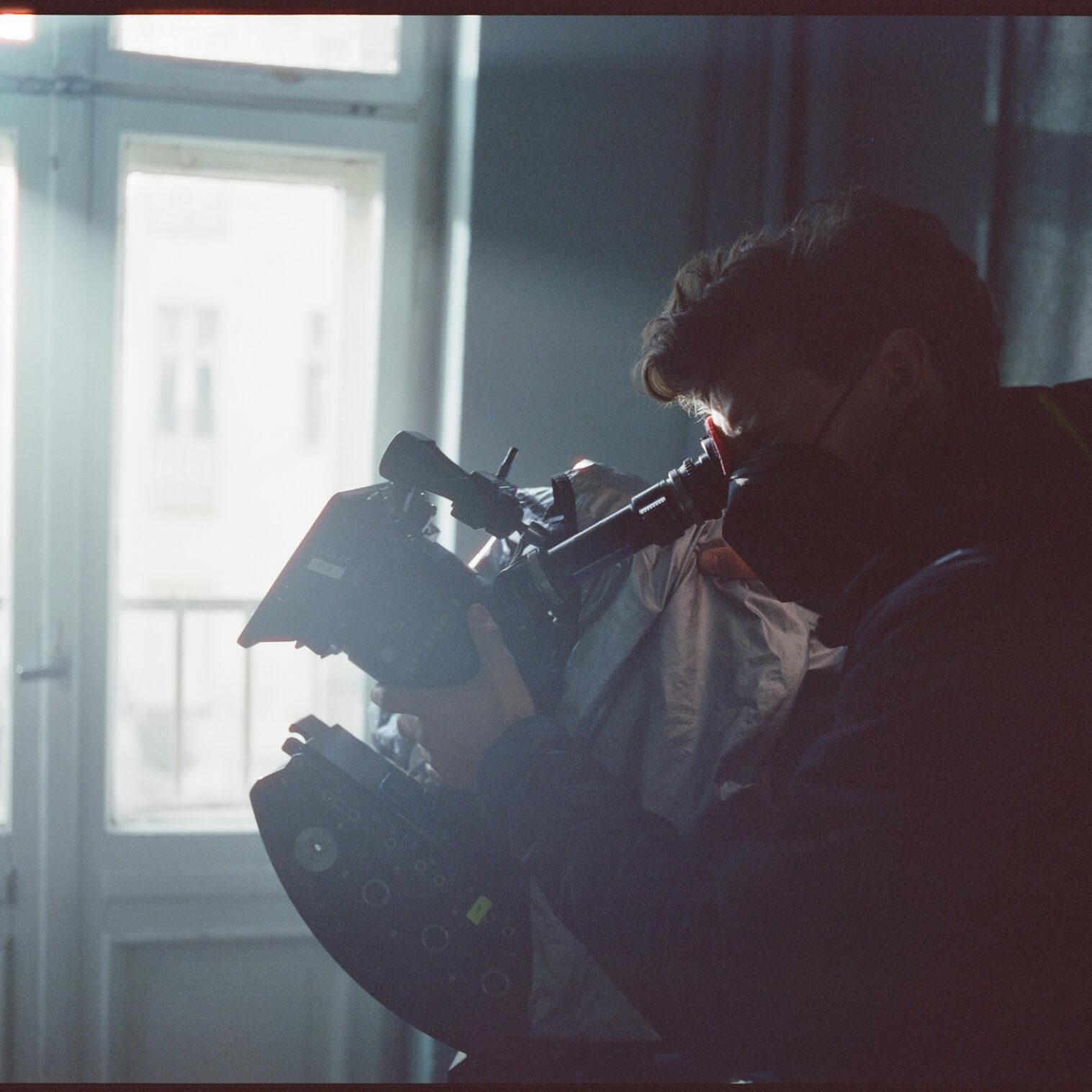 A person wearing a mask operates a professional video camera equipped with a ZEISS lens in a softly lit indoor setting.
