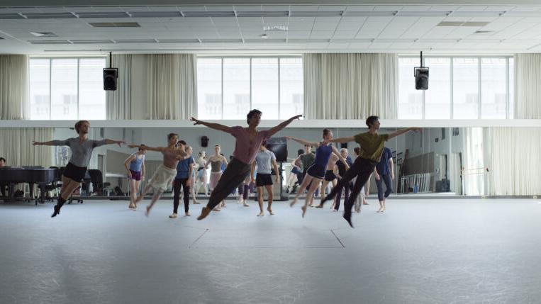 A group of dancers rehearses in a spacious, well-lit studio with large windows, as others observe in the background.