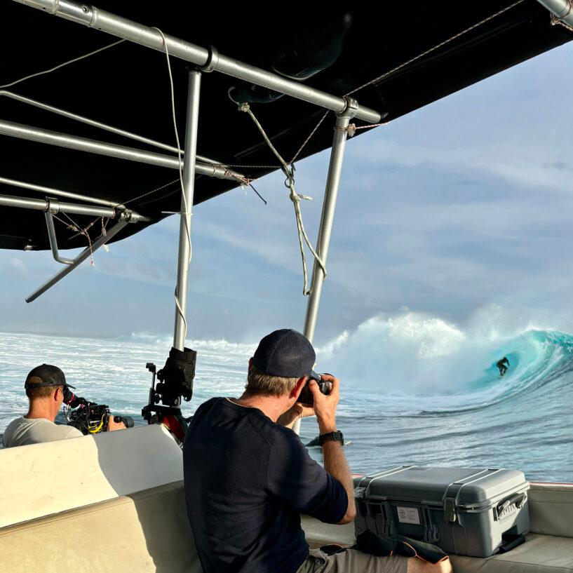Two photographers on a boat capture a surfer riding a large wave using professional ZEISS camera lenses and equipment.