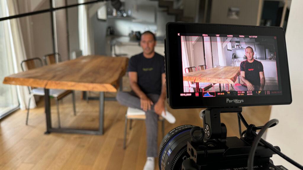 A man is being filmed in a modern, well-lit room with a ZEISS camera setup, and he appears on the camera monitor in front.