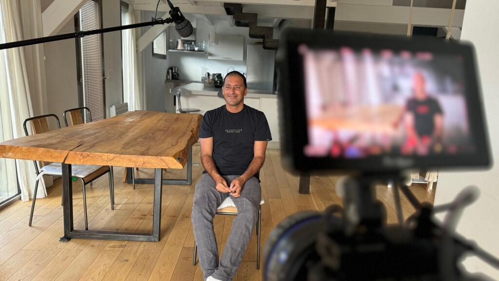 A man sits for an interview in a modern, naturally lit room with a wooden table, while being filmed by a professional camera setup with a visible monitor and boom microphone, showcasing ZEISS video production equipment.