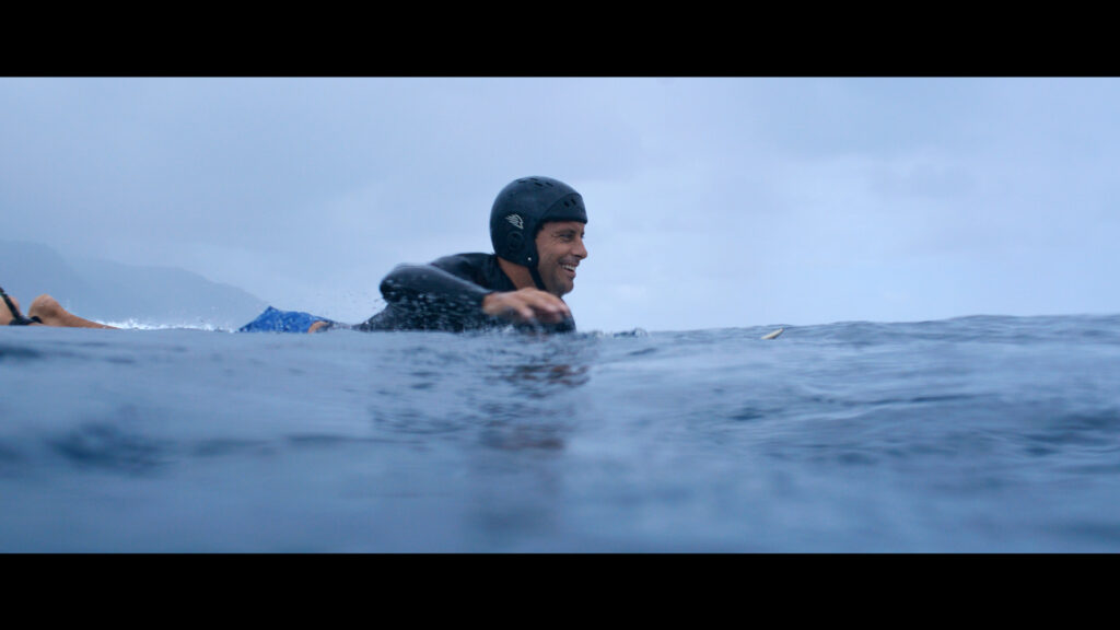 A man in a wetsuit and helmet is paddling on a surfboard in the ocean under a cloudy sky.