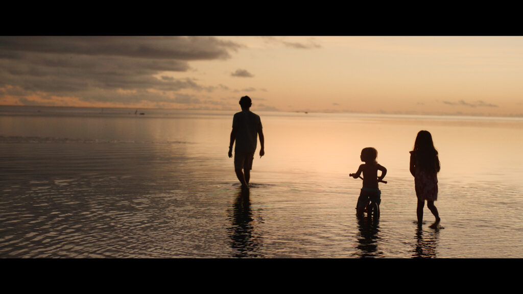 Three people, including a child on a tricycle, walk through shallow water during a calm sunset.