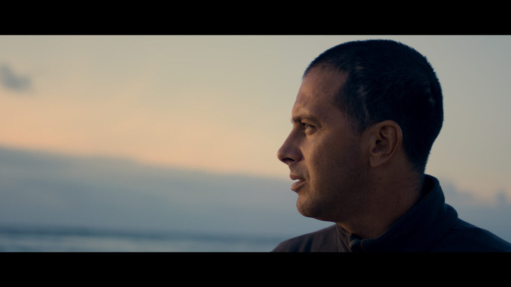 A man with short hair looks thoughtfully into the distance during sunset by the water, with soft lighting highlighting his profile.