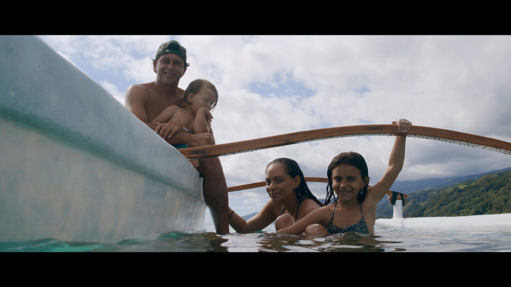 A family of four enjoys time in the water beside a wooden outrigger canoe under a partly cloudy sky.