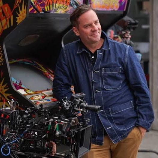 A man in a denim jacket stands next to an arcade machine and a professional cinema camera rig equipped with ZEISS lenses.
