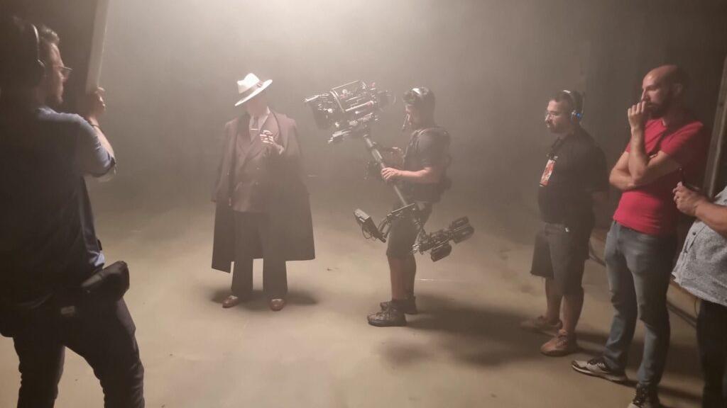 A film crew captures a dramatic scene with a smoke-filled set, featuring a camera operator using a stabilizer rig and an actor in a trench coat and hat, during a production shoot with ZEISS cinema lenses.
