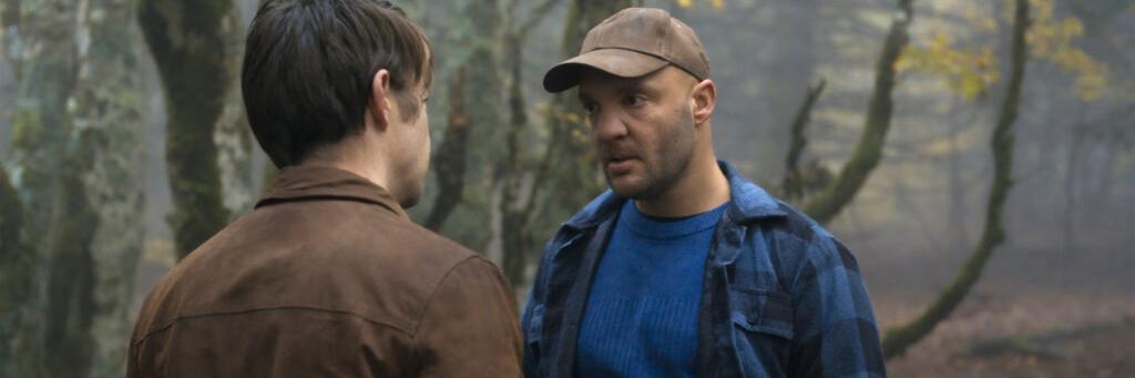 Two men are standing in a foggy forest having a serious conversation, with autumn trees in the background.