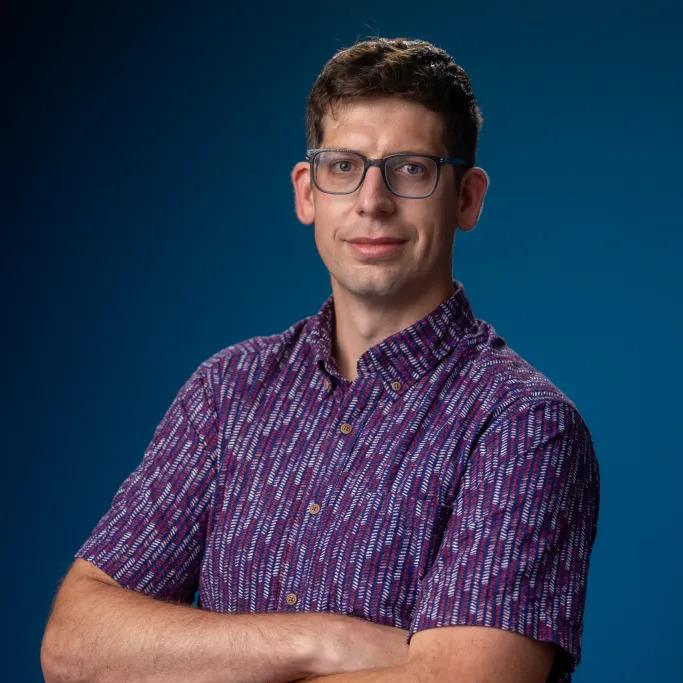 A man wearing glasses and a patterned short-sleeve shirt stands with arms crossed in front of a blue background.