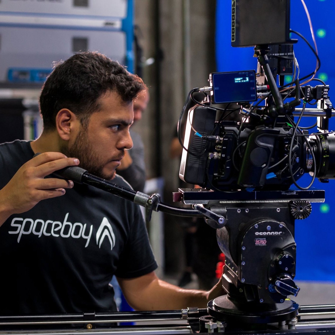 A cameraman operates a professional cinema camera mounted on a dolly system, equipped with ZEISS lenses, in a studio setting.
