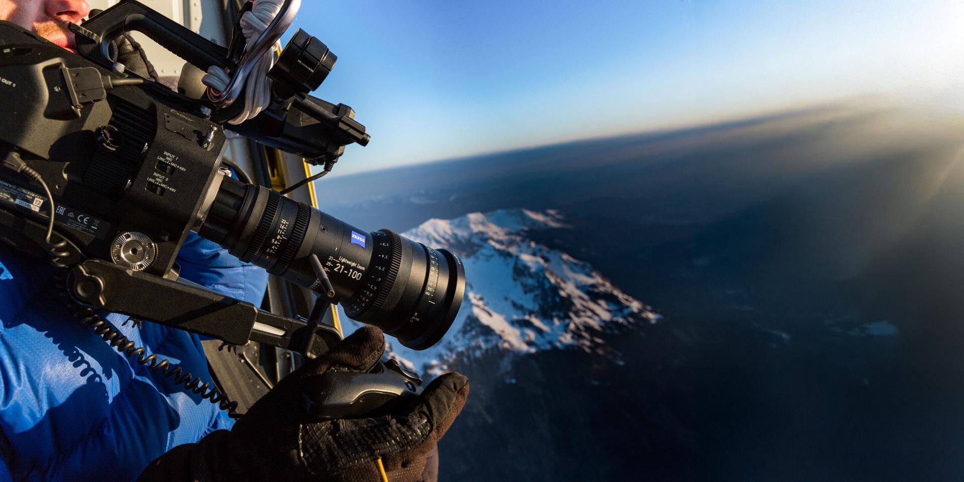 A professional camera equipped with a ZEISS lens is being operated from a helicopter to film a snow-covered mountain landscape.