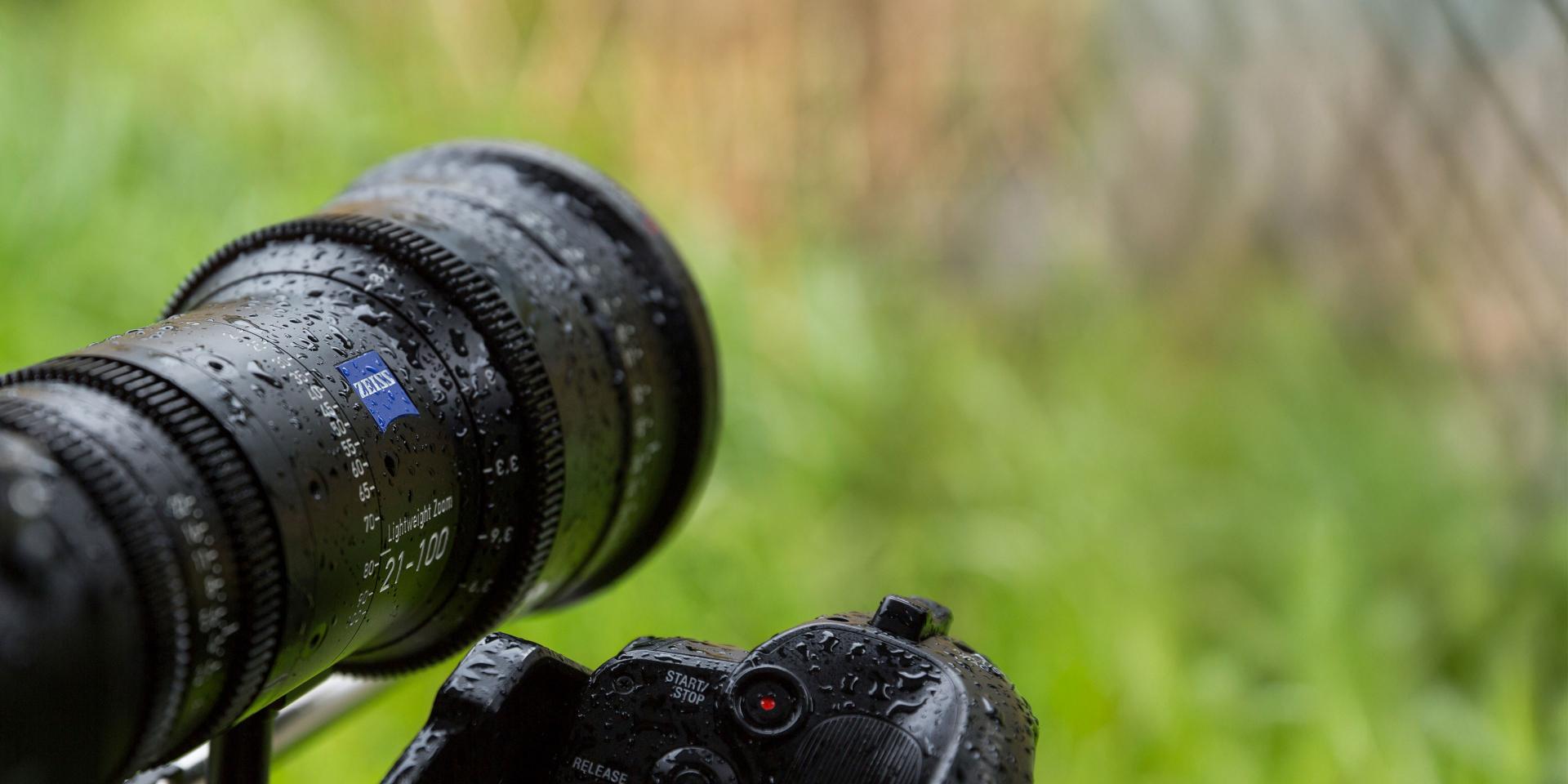 Close-up of a professional camera equipped with a ZEISS lens, covered in water droplets, set against a blurred green outdoor background.