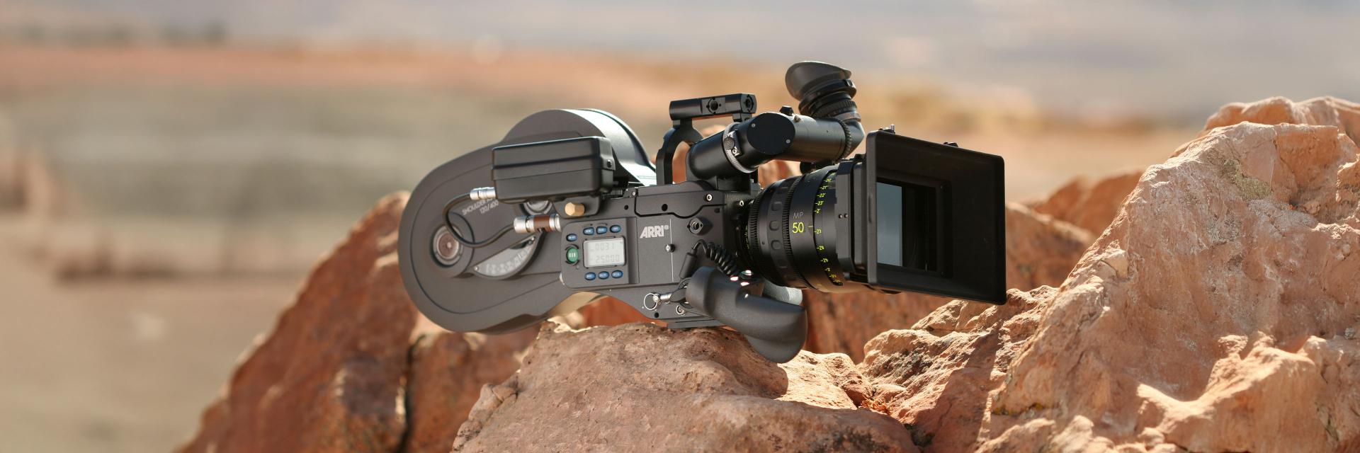 Professional film camera equipped with a ZEISS lens positioned on rocky terrain in a desert landscape.