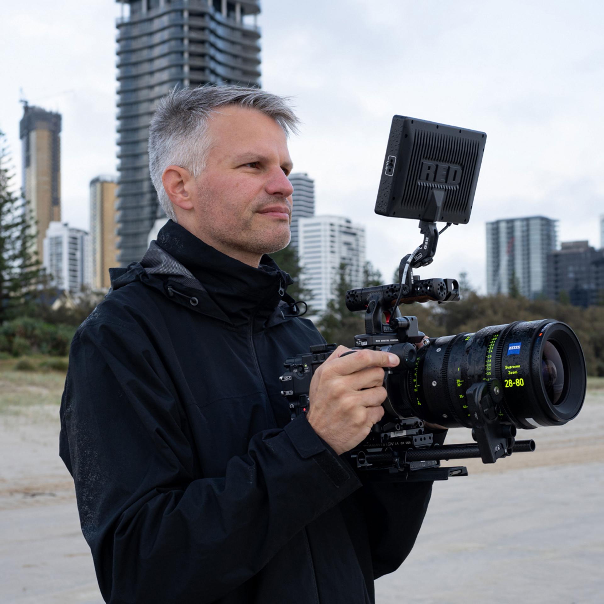 A man holds a professional cinema camera equipped with a ZEISS lens, filming outdoors near tall buildings and trees.