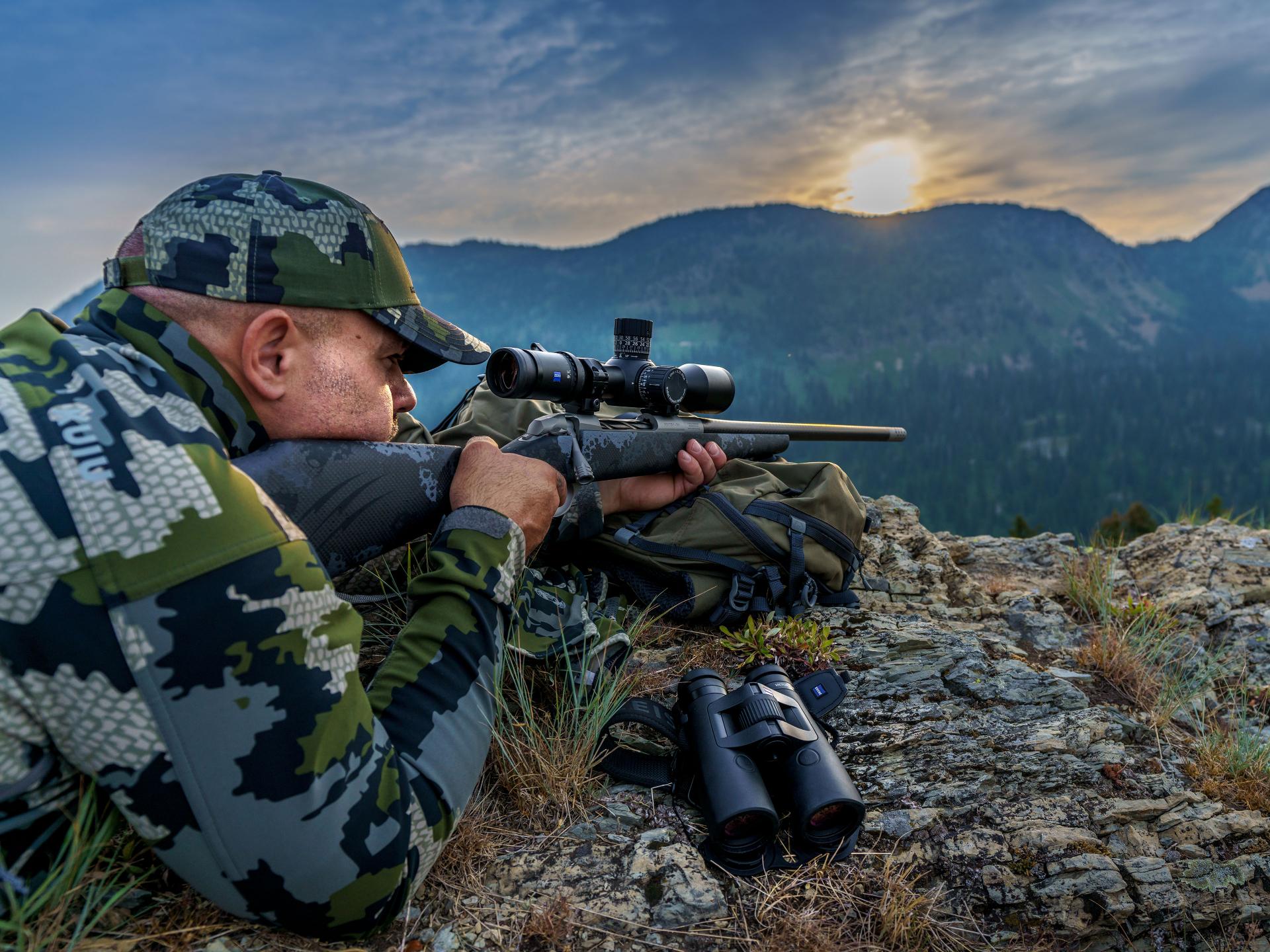 A hunter lies on rocky terrain using a ZEISS LRP S5 riflescope for long-range accuracy, with binoculars beside him and mountains in the background.
