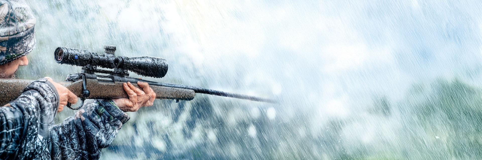 A hunter takes careful aim with his rifle equipped with a ZEISS riflescope while standing in heavy rain, demonstrating reliability and precision even in extreme weather conditions.