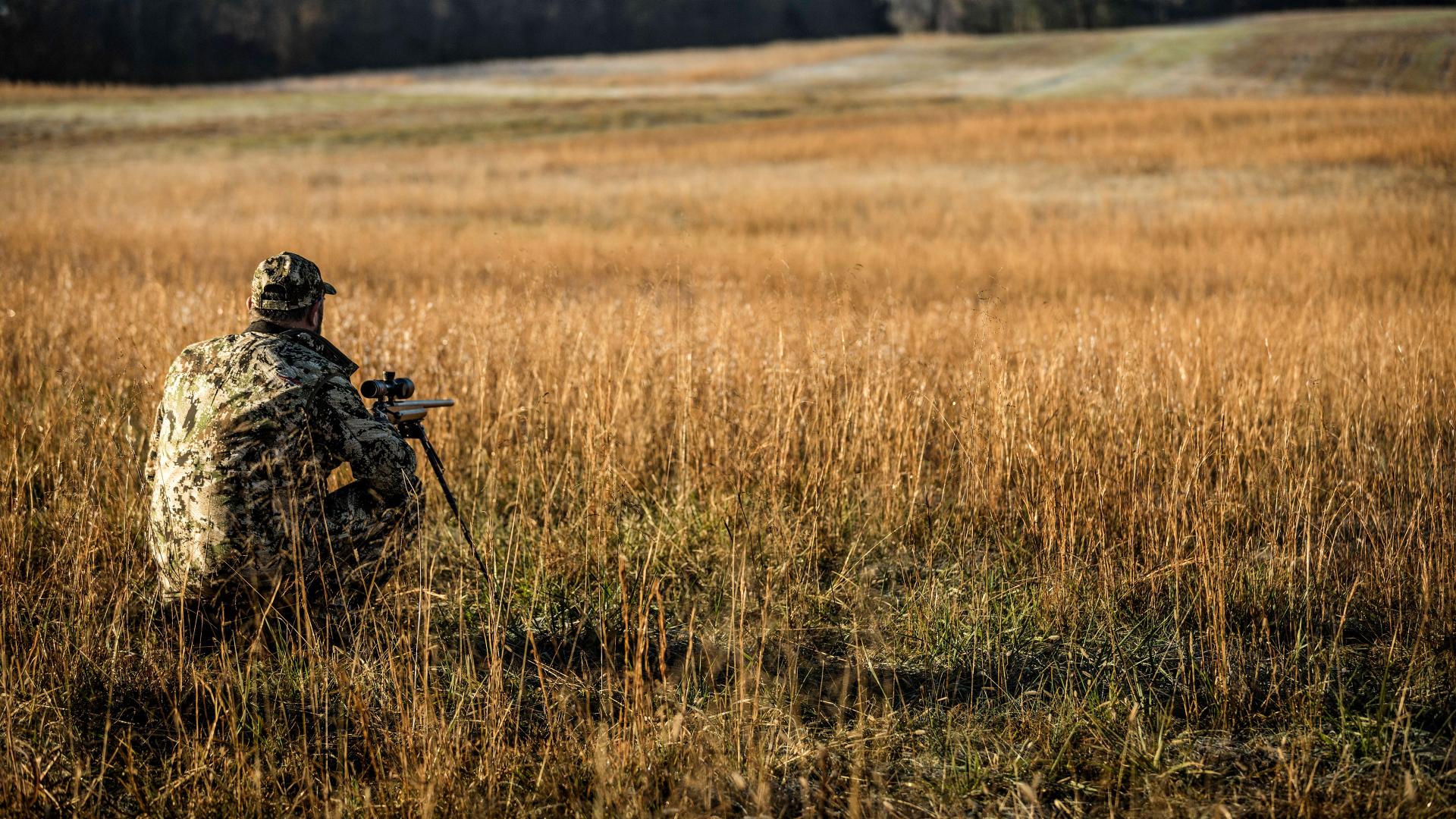 Hunter in camouflage aiming across an open field, showcasing ZEISS riflescope’s 90% light transmission for clear visibility in low light