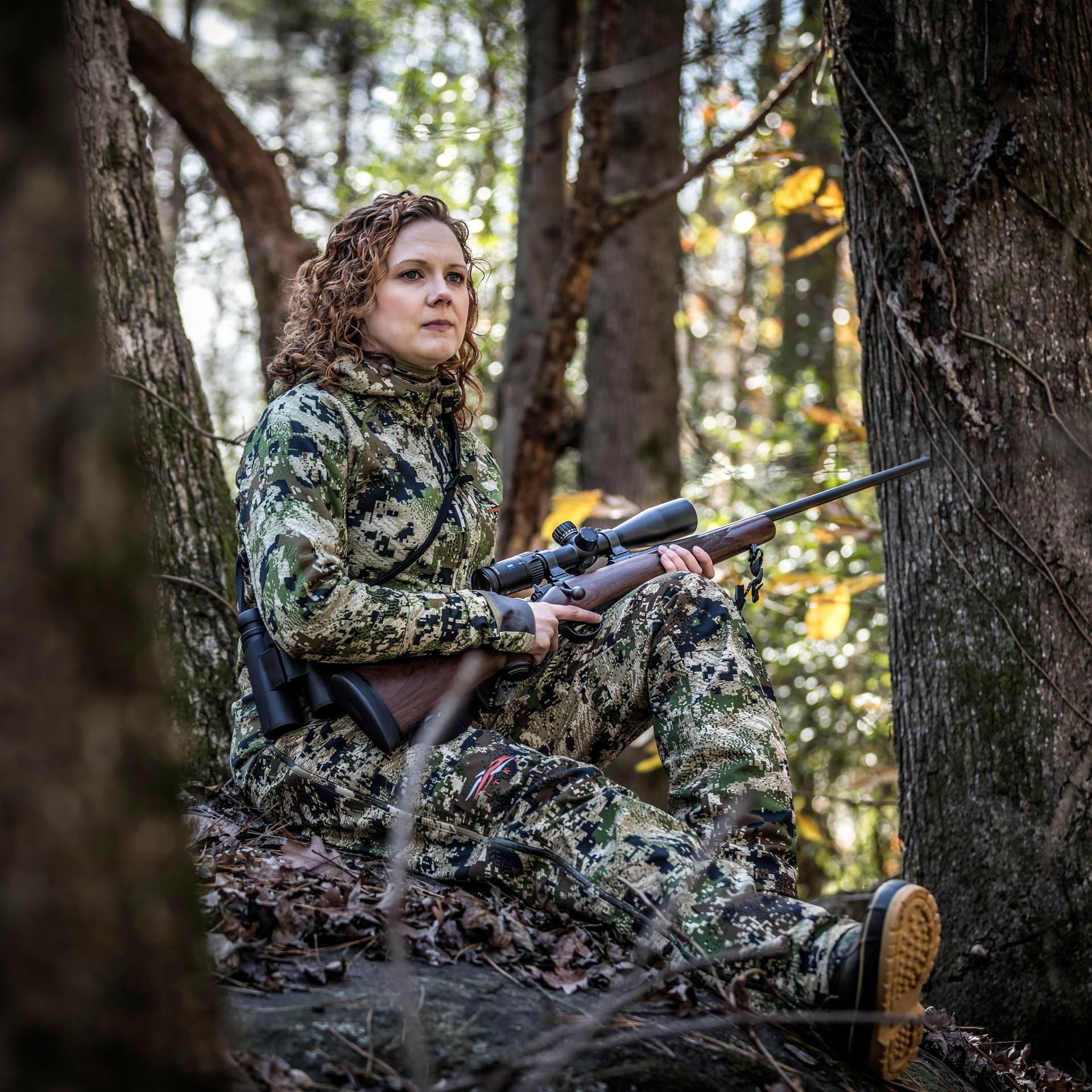 Female hunter in camouflage sitting between trees with a rifle equipped with a ZEISS Conquest V4 4-16x44 riflescope, ideal for lightweight precision in mountain hunts