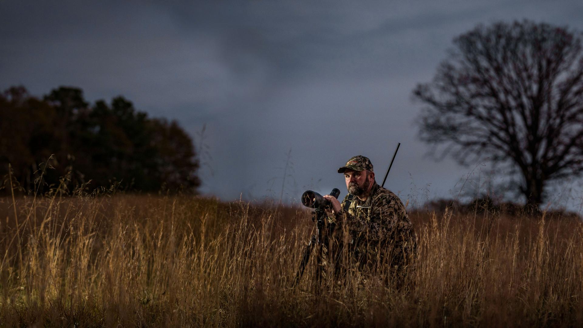 A man in camouflage gear uses a ZEISS spotting scope on a tripod in a tall grassy field at dusk.