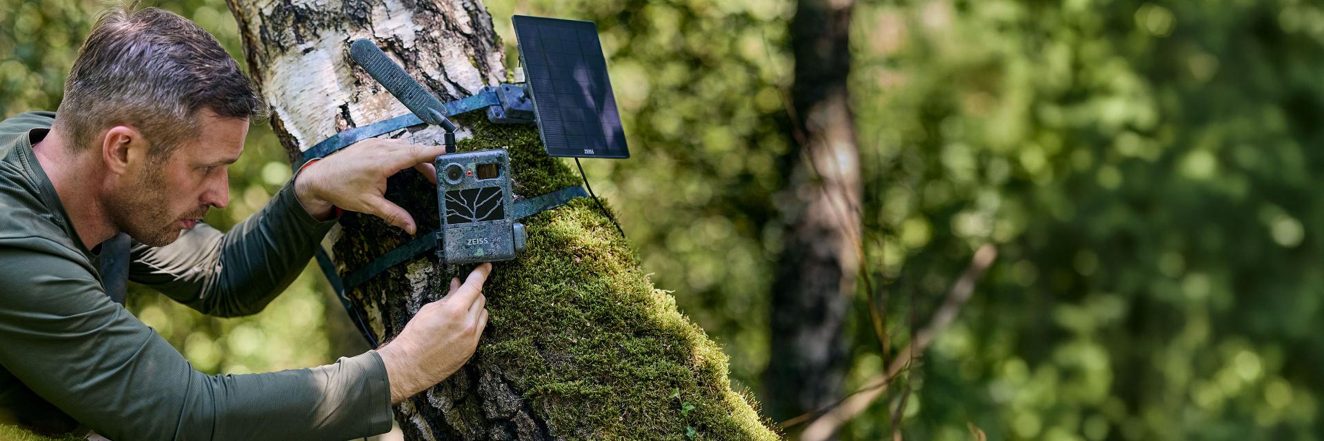 A man is mounting a trail camera and a solar panel to a moss-covered tree in a forest setting for ZEISS.
