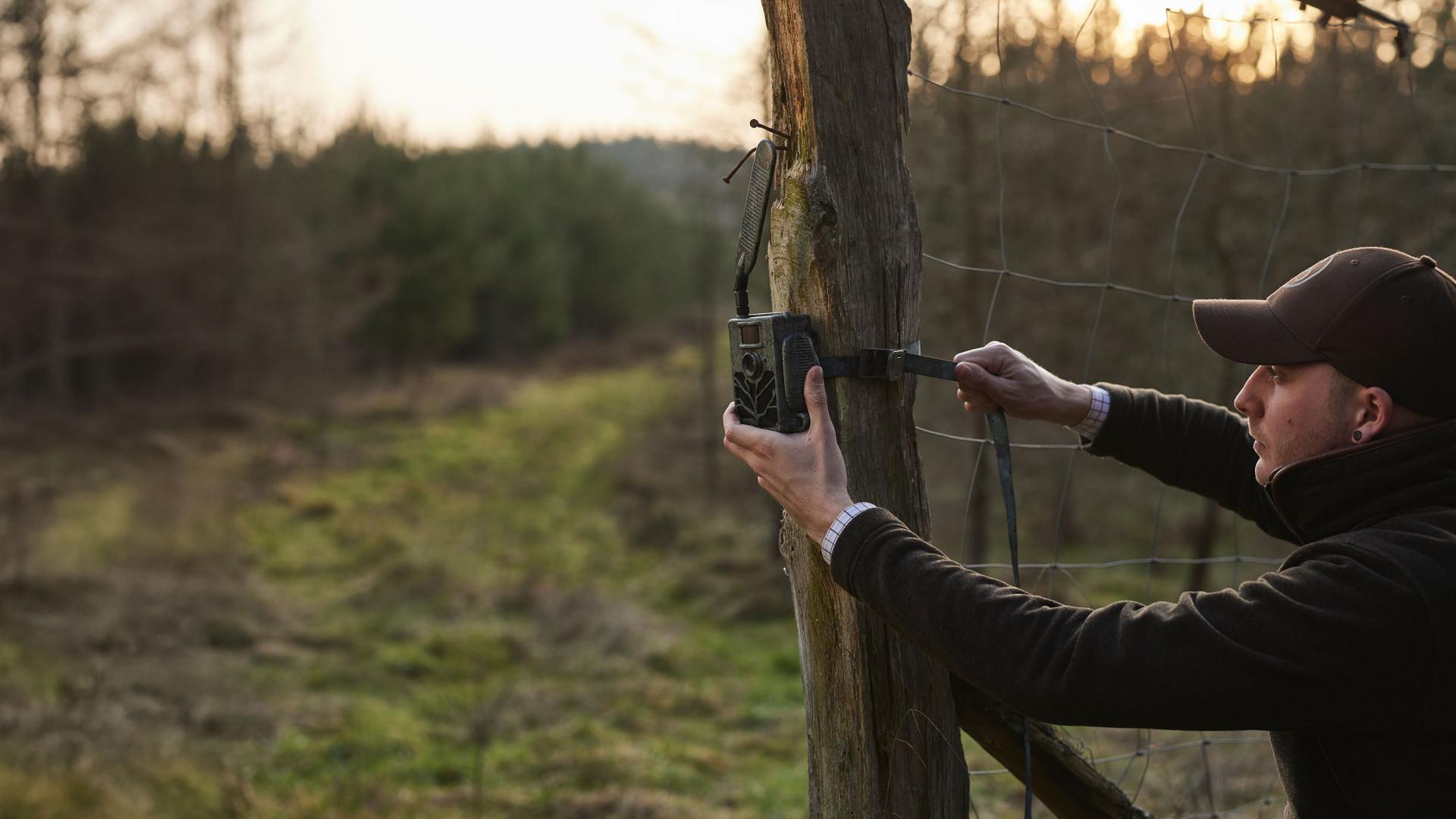 A man in outdoor clothing mounts a ZEISS trail camera to a wooden fence post in a forested area at sunset.