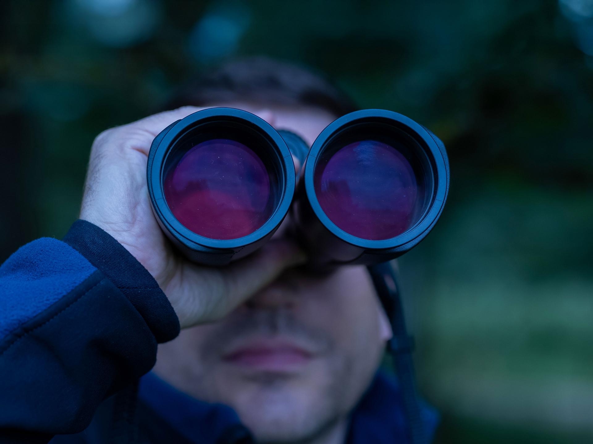 A person holding and looking through a pair of ZEISS binoculars with red-tinted lenses in an outdoor setting.
