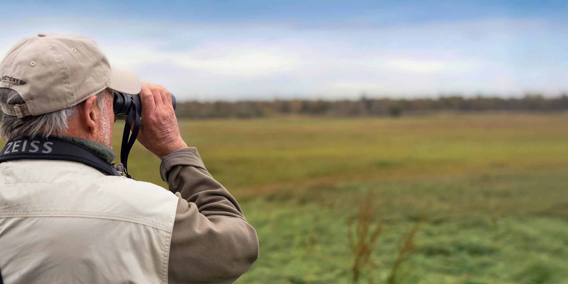 An older man wearing a ZEISS-branded strap uses binoculars to observe a wide open grassy landscape.