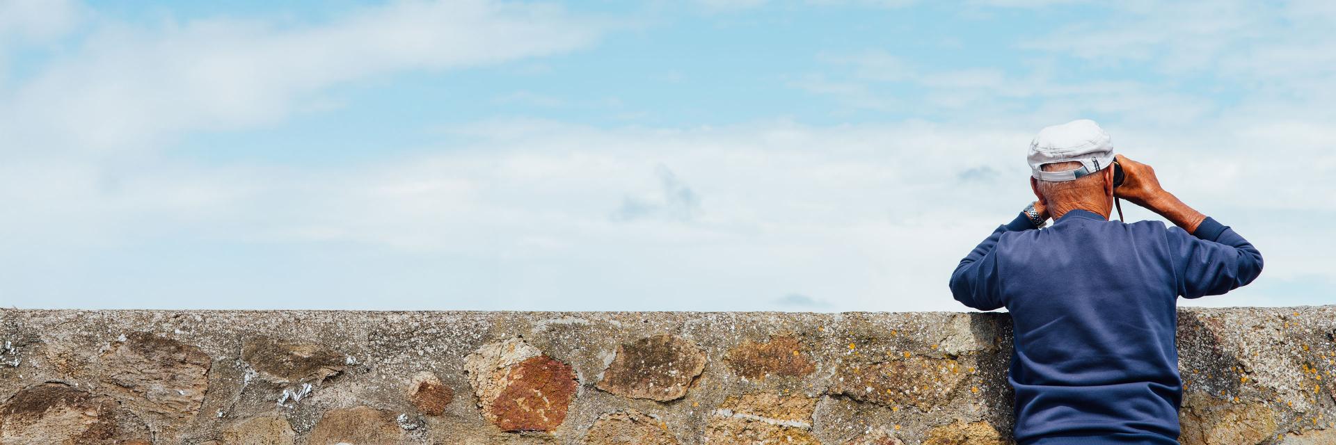 An older man wearing a blue sweater and white cap looks through binoculars over a stone wall toward the sky.