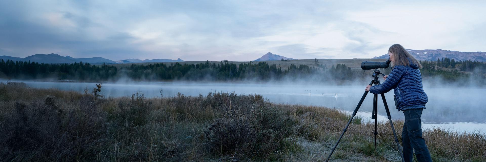 A person using a ZEISS spotting scope on a tripod to observe a misty lakeside landscape during early morning.
