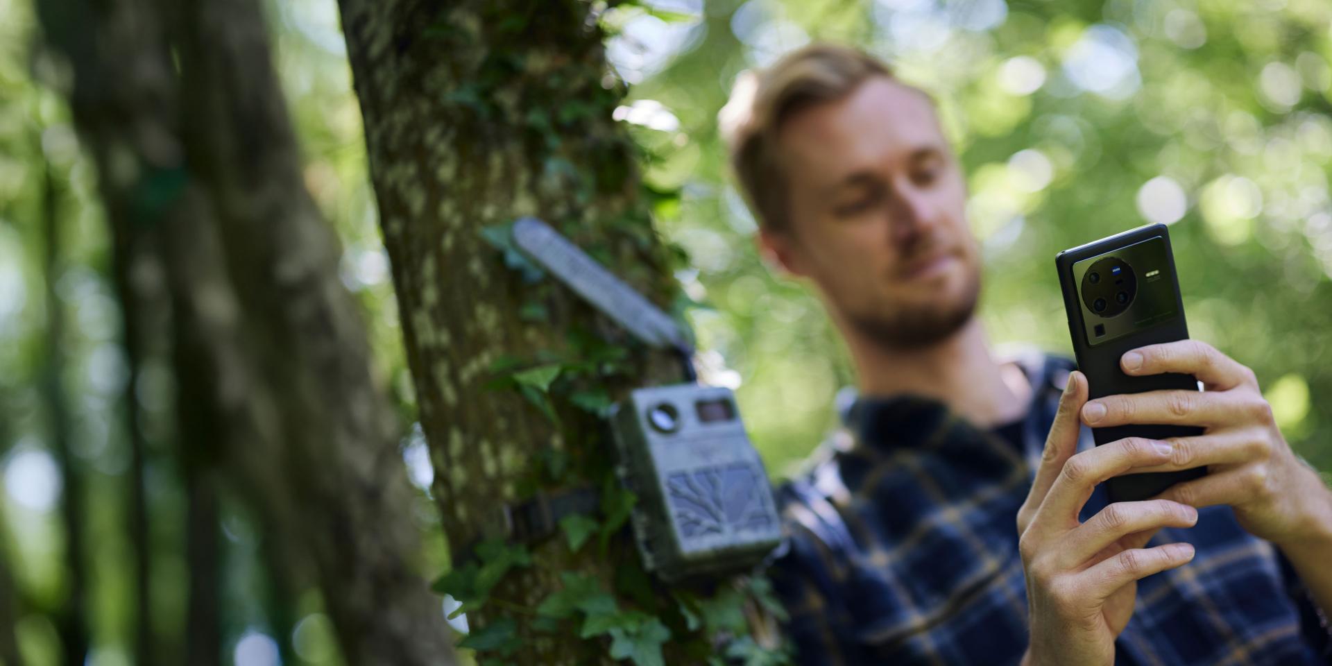 A man uses a smartphone equipped with a ZEISS lens near a tree-mounted wildlife camera in a forest setting.