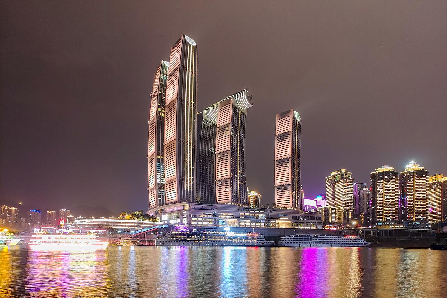 Modern high-rise buildings illuminated at night are reflected in the calm water of a river, showcasing vibrant city lights and architectural detail captured with ZEISS clarity.