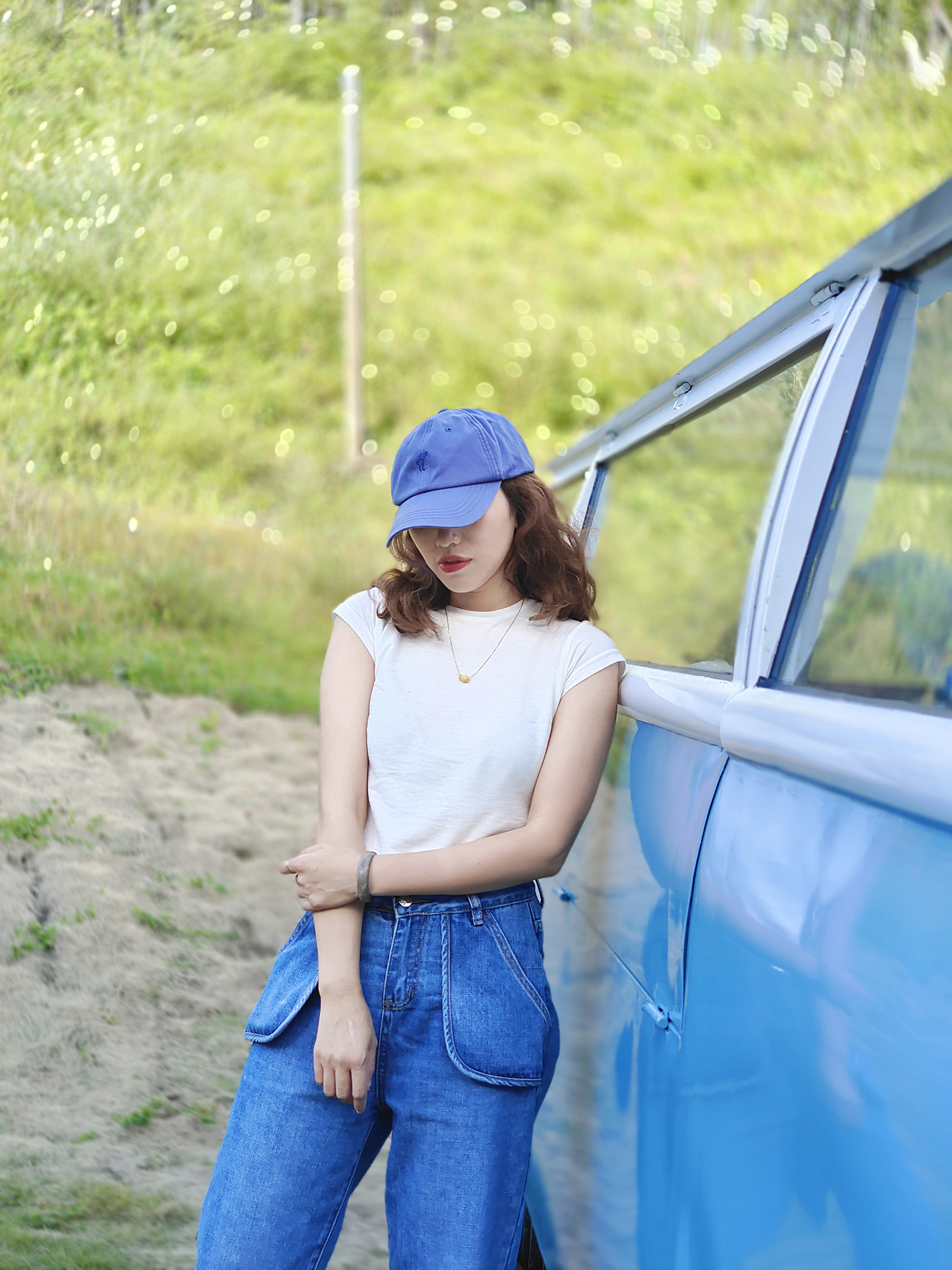 A woman wearing a blue cap and white shirt stands beside a blue vehicle in a grassy outdoor setting, captured in sharp detail with a ZEISS lens.