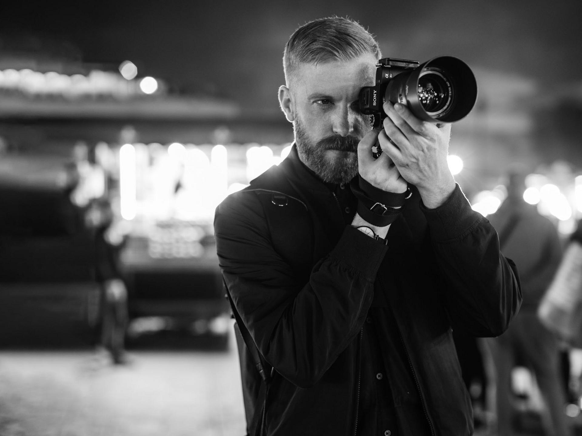 A man is photographing a night scene using a camera equipped with a ZEISS lens.