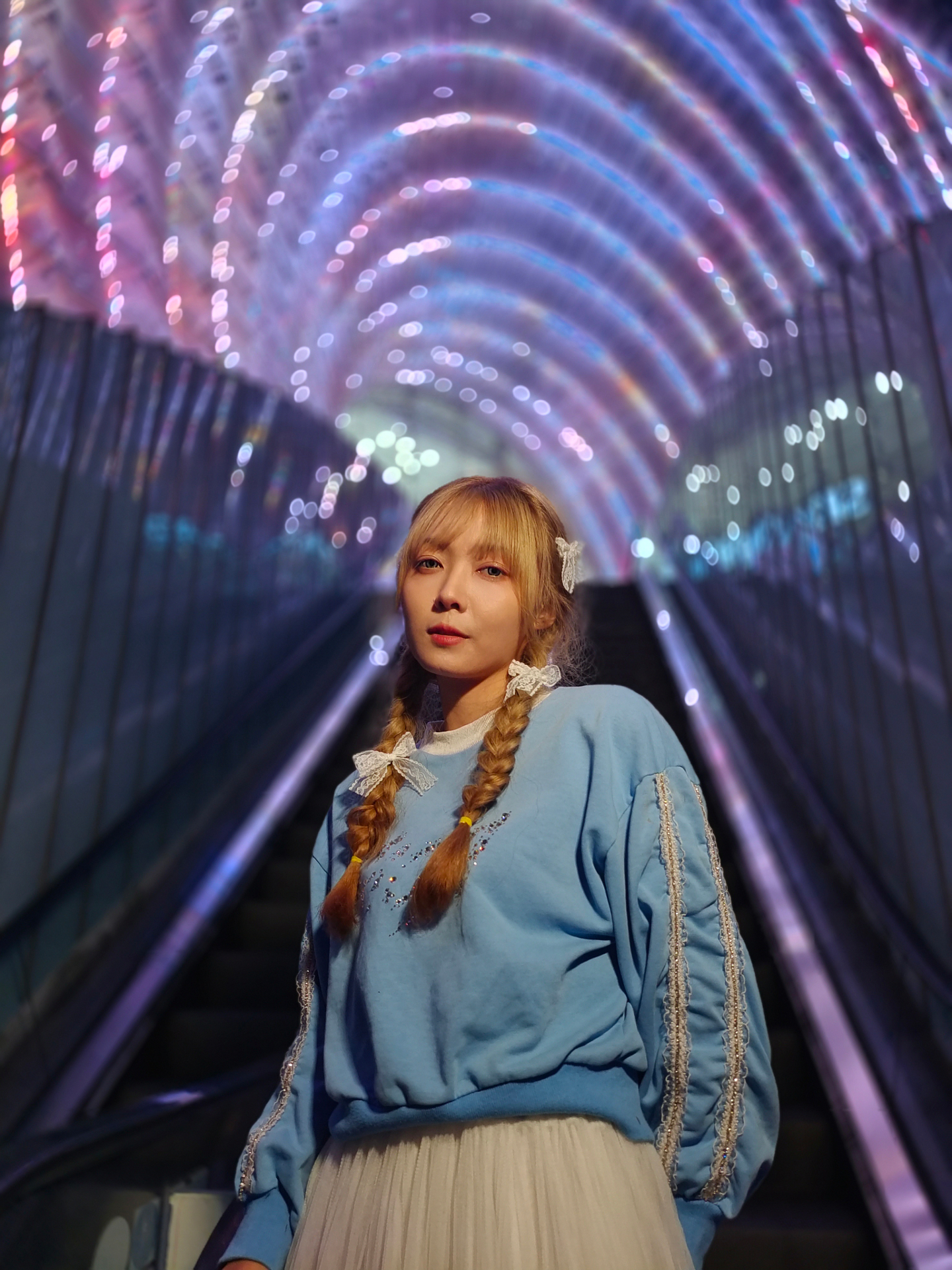 A woman with braided hair stands on an escalator beneath a tunnel of colorful, glowing lights, captured with sharp detail and vibrant colors.