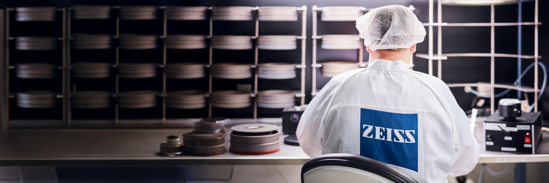 Technician in a cleanroom environment wearing a ZEISS lab coat and hairnet, working with optical equipment and materials.