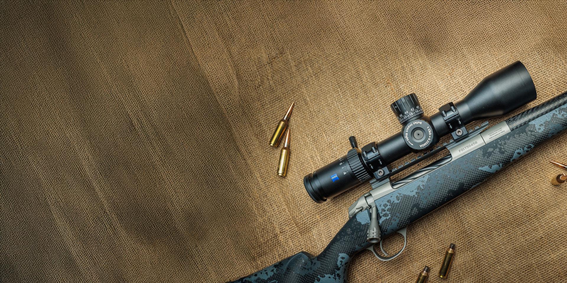A precision rifle with a mounted ZEISS riflescope is displayed on a textured fabric surface alongside several rifle cartridges.