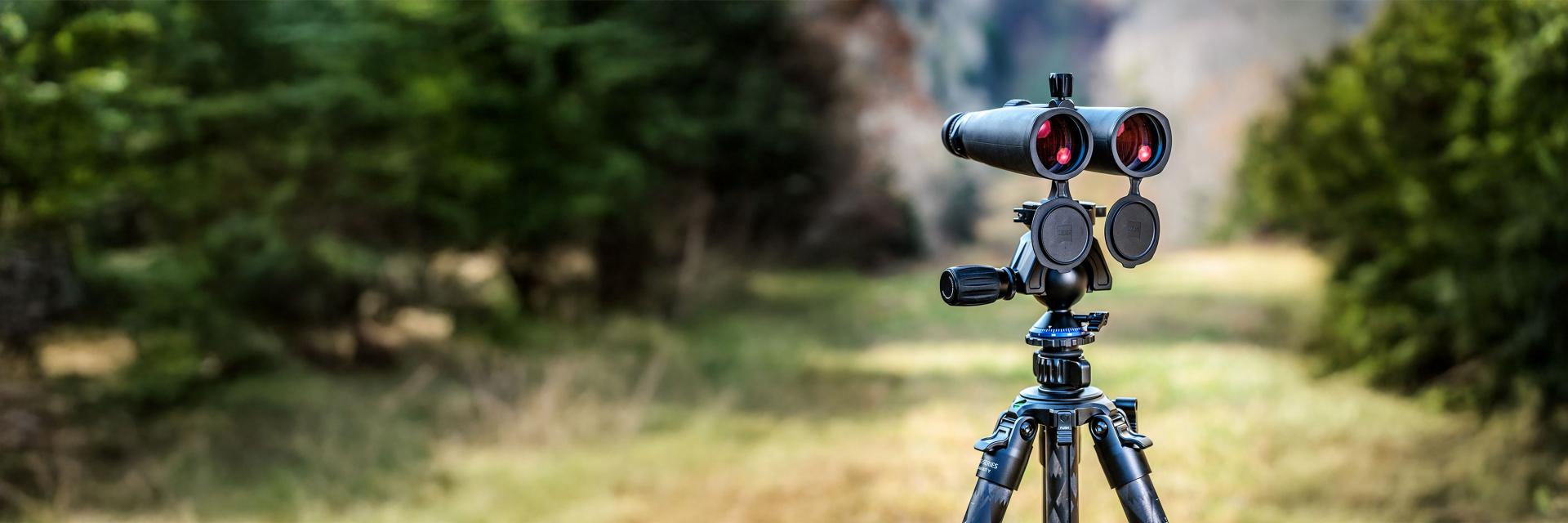 A pair of ZEISS binoculars mounted on a tripod, set against a blurred outdoor background at sunset.