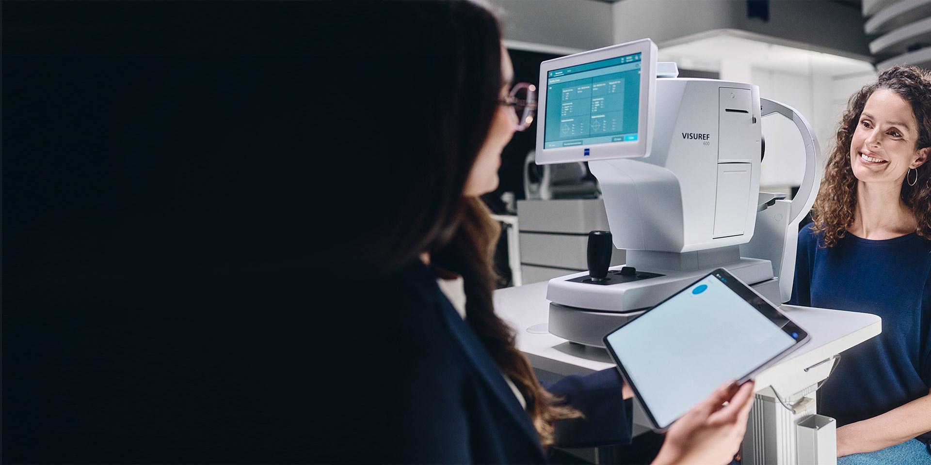 Un&apos;ottico donna che guarda il suo tablet seduta davanti a ZEISS VISUREF 600 e parla con il cliente.