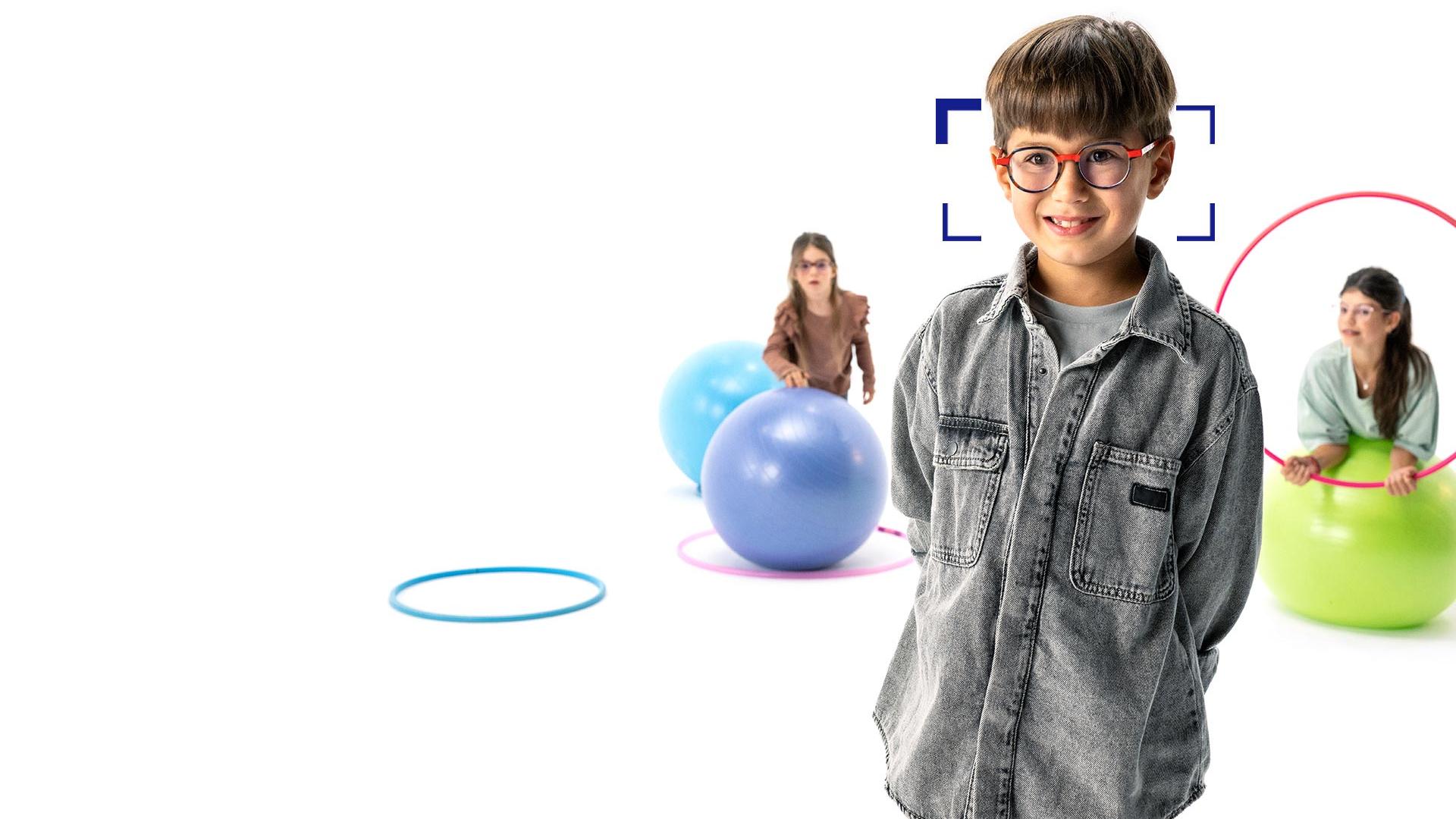Bambino con i capelli marroni e gli occhiali tondi che indossa le lenti ZEISS MyoCare in piedi in primo piano sorride all’obiettivo. Sullo sfondo due bambine che indossano le lenti ZEISS MyoCare giocano con l’hula hoop e la palla.