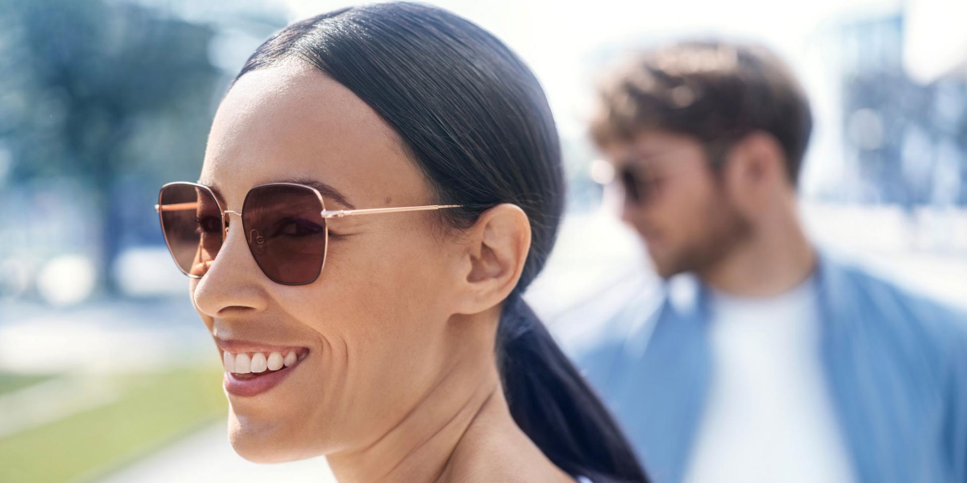 Davanti: donna sorridente con occhiali da sole, sullo sfondo: un uomo con occhiali