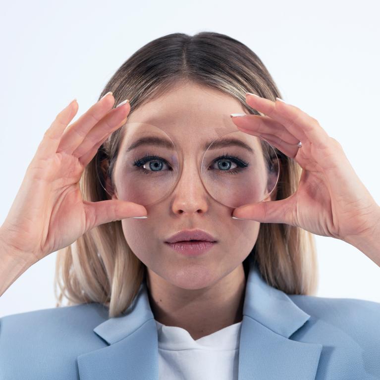 Giovane donna bionda che tiene in mano di fronte agli occhi delle lenti per mostrare il bel look privo di effetti comici grazie alle lenti monofocali ZEISS ClearView.