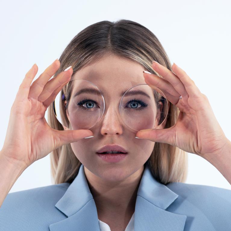 Giovane donna bionda che tiene in mano di fronte agli occhi delle lenti per mostrare l&apos;&quot;effetto tiny-eye&quot; causato da occhiali negativi spessi.