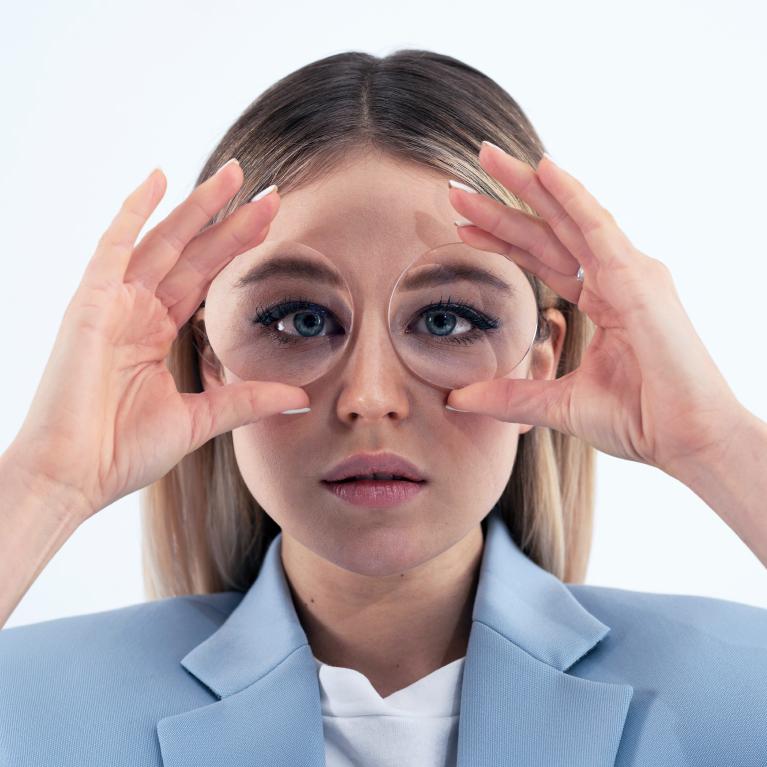 Giovane donna bionda che tiene in mano di fronte agli occhi delle lenti per mostrare l&apos;&quot;effetto fisheye&quot; causato da occhiali spessi.