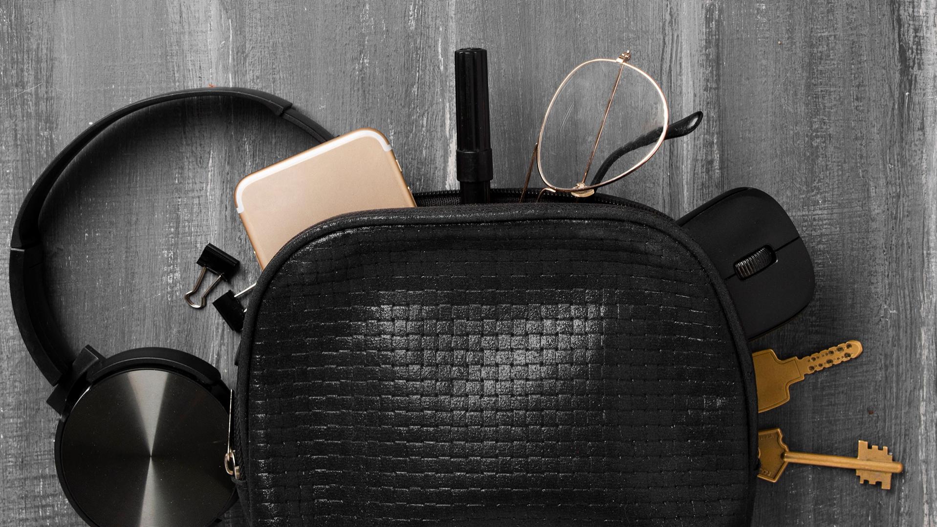 Una borsetta nera da cui sono scivolati fuori cuffie, cellulare, chiavi, graffette, penna, mouse per PC e occhiali con lenti ZEISS dotate di trattamenti DuraVision, che ora giacciono per metà sul pavimento grigio.