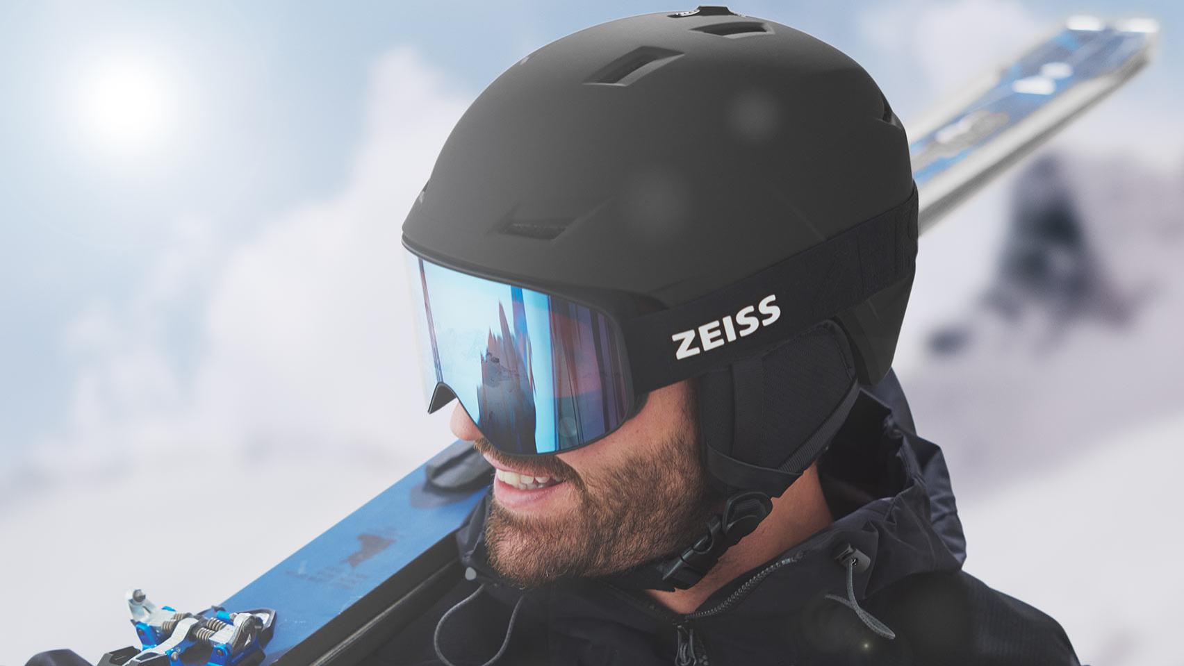 Un uomo indossa un casco nero con maschera da sci ZEISS blu a specchio e porta gli sci sulla spalla destra.