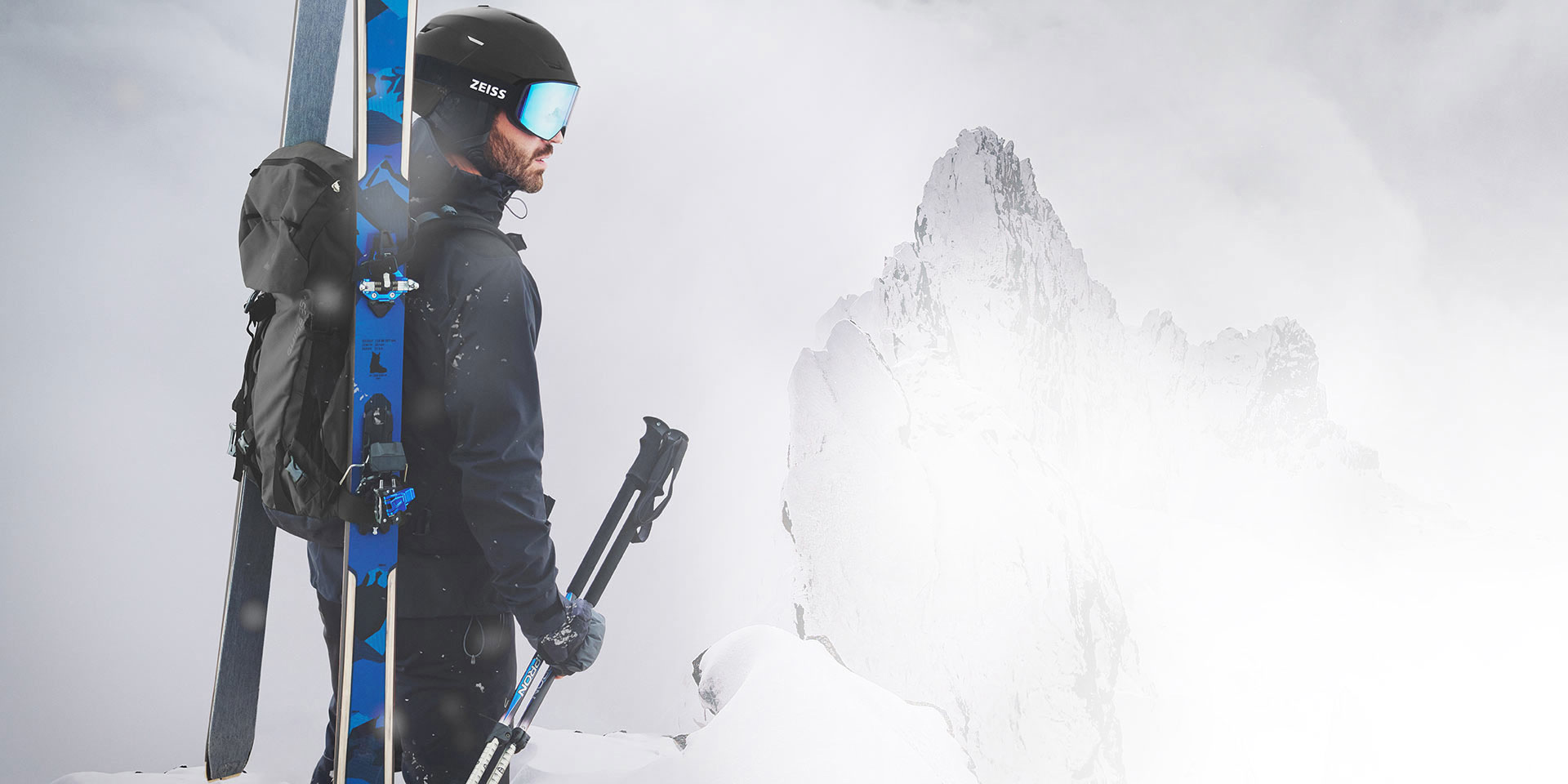 Un uomo indossa un casco nero con maschera da sci ZEISS blu a specchio, porta gli sci e uno zaino sulla schiena e tiene i bastoncini nella mano destra. È in piedi sulla cima di una montagna.