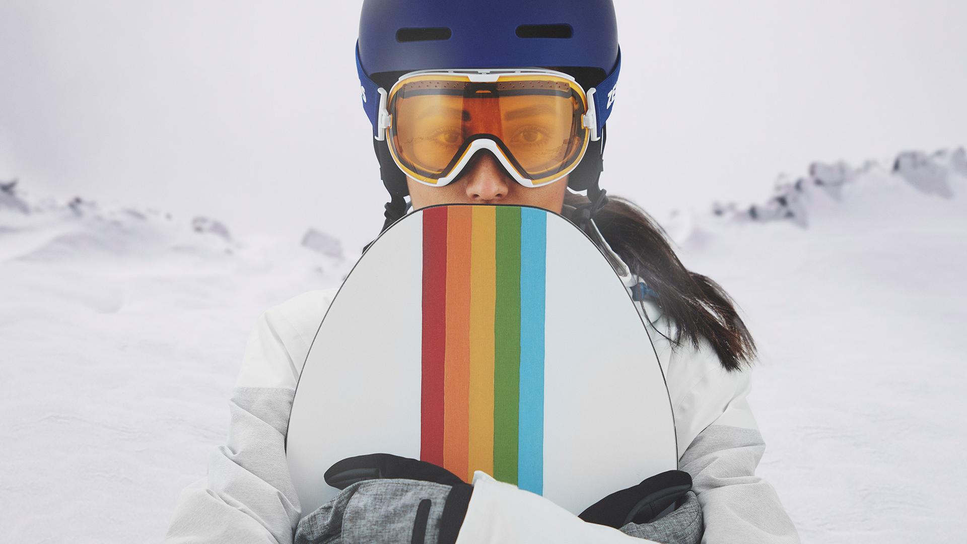 Una giovane donna indossa una maschera da sci con tecnologia ZEISS Interchangeable e tiene in mano uno snowboard.
