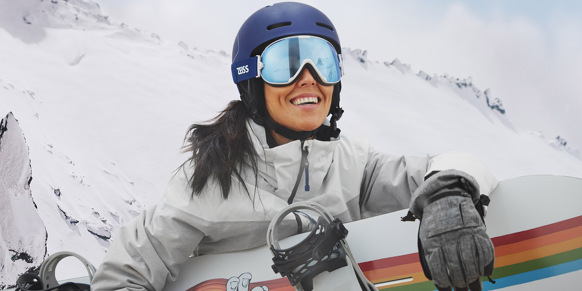 Una giovane donna indossa maschere da sci con tecnologia ZEISS Interchangeable e tiene in mano uno snowboard.