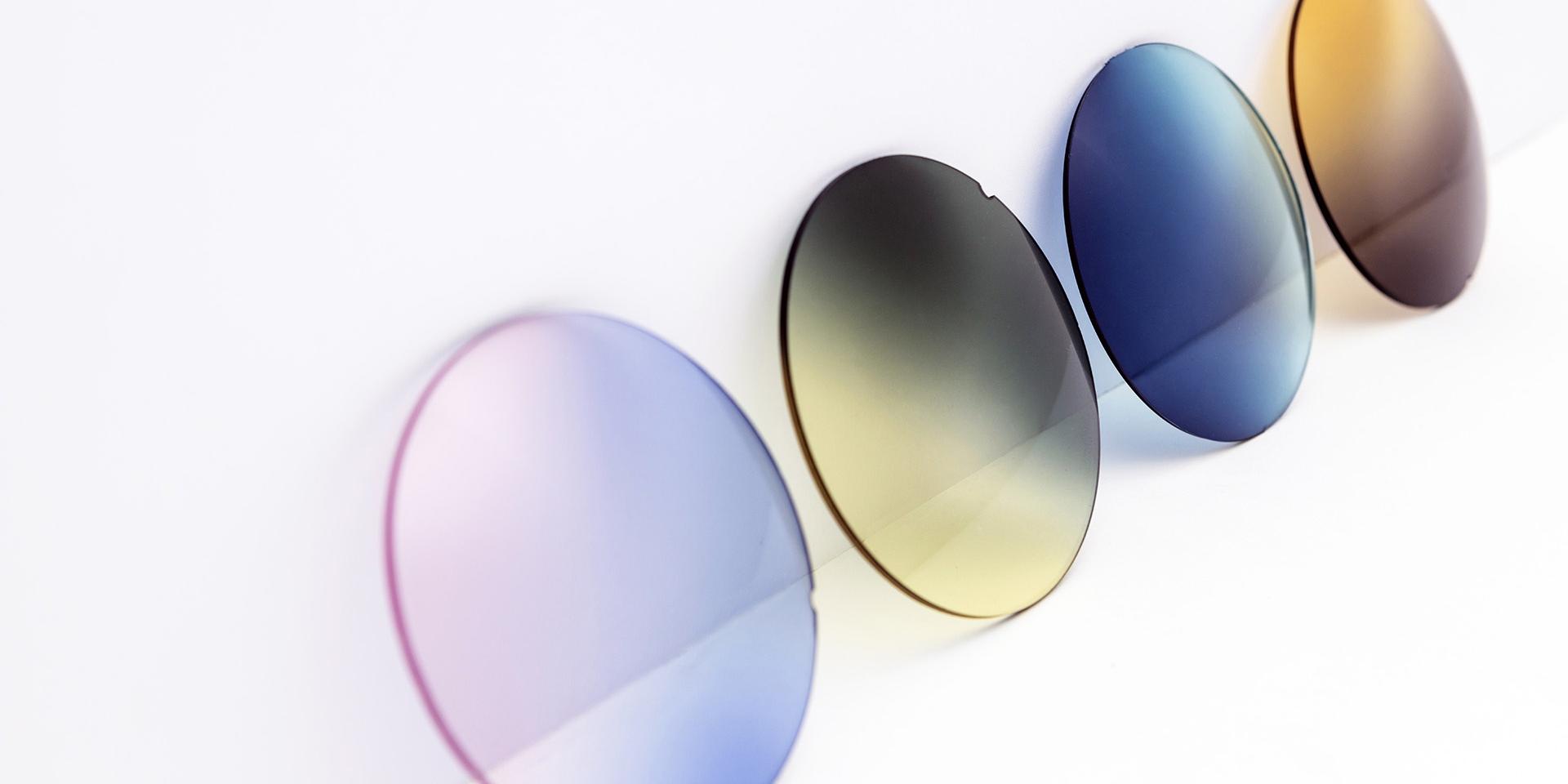 Lenti per occhiali da sole di diversi colori appoggiate su una superficie bianca: sfumature rosa-viola, giallo-grigio, blu e marrone.